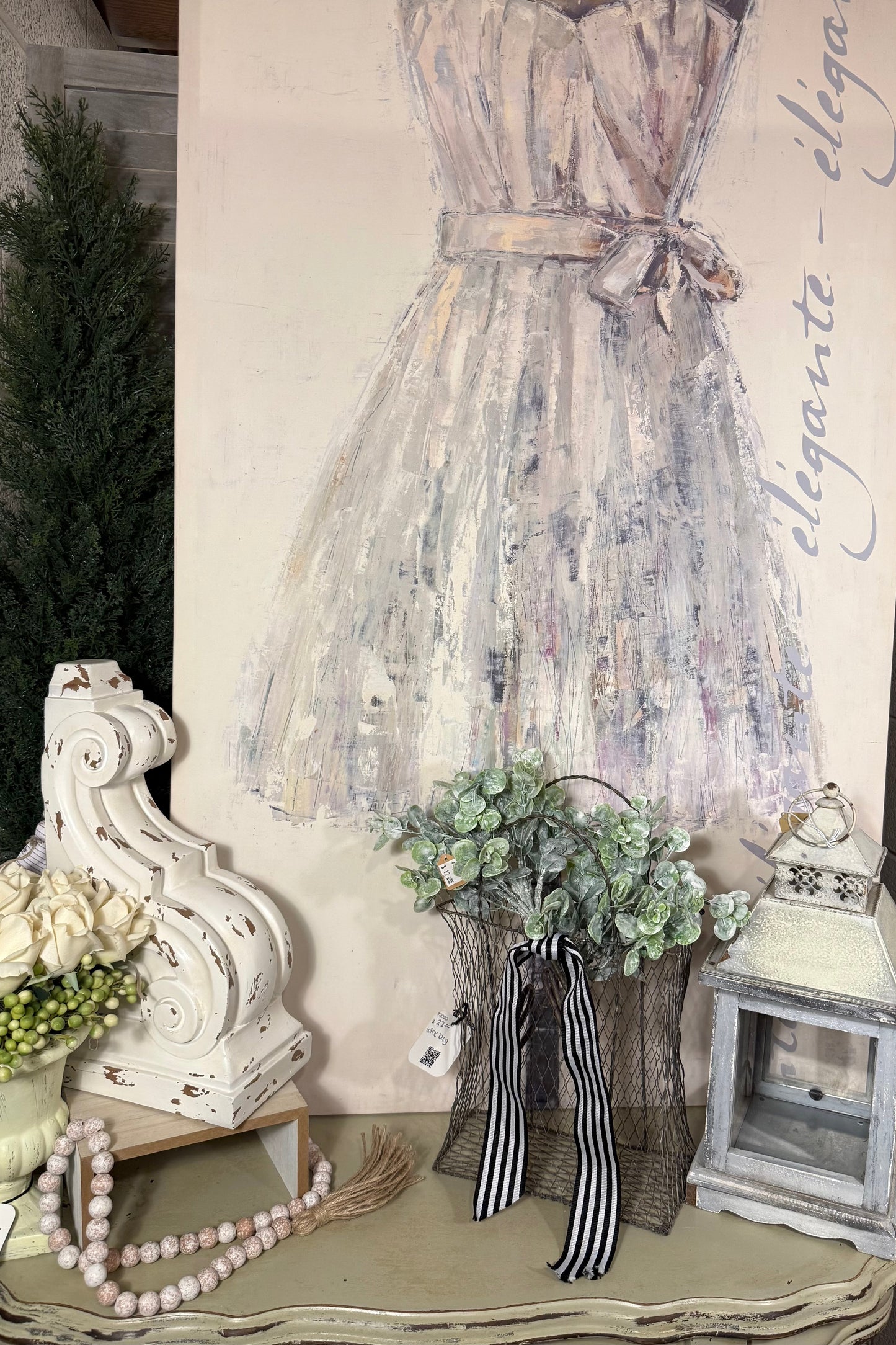 Chic feminine décor setup featuring a pastel dress canvas with 'elegante' script, distressed white corbel, faux white roses, vintage beaded garland, wire basket with greenery and striped ribbon, and a rustic lantern. Perfect for shabby chic, French country, or boutique-style home accents