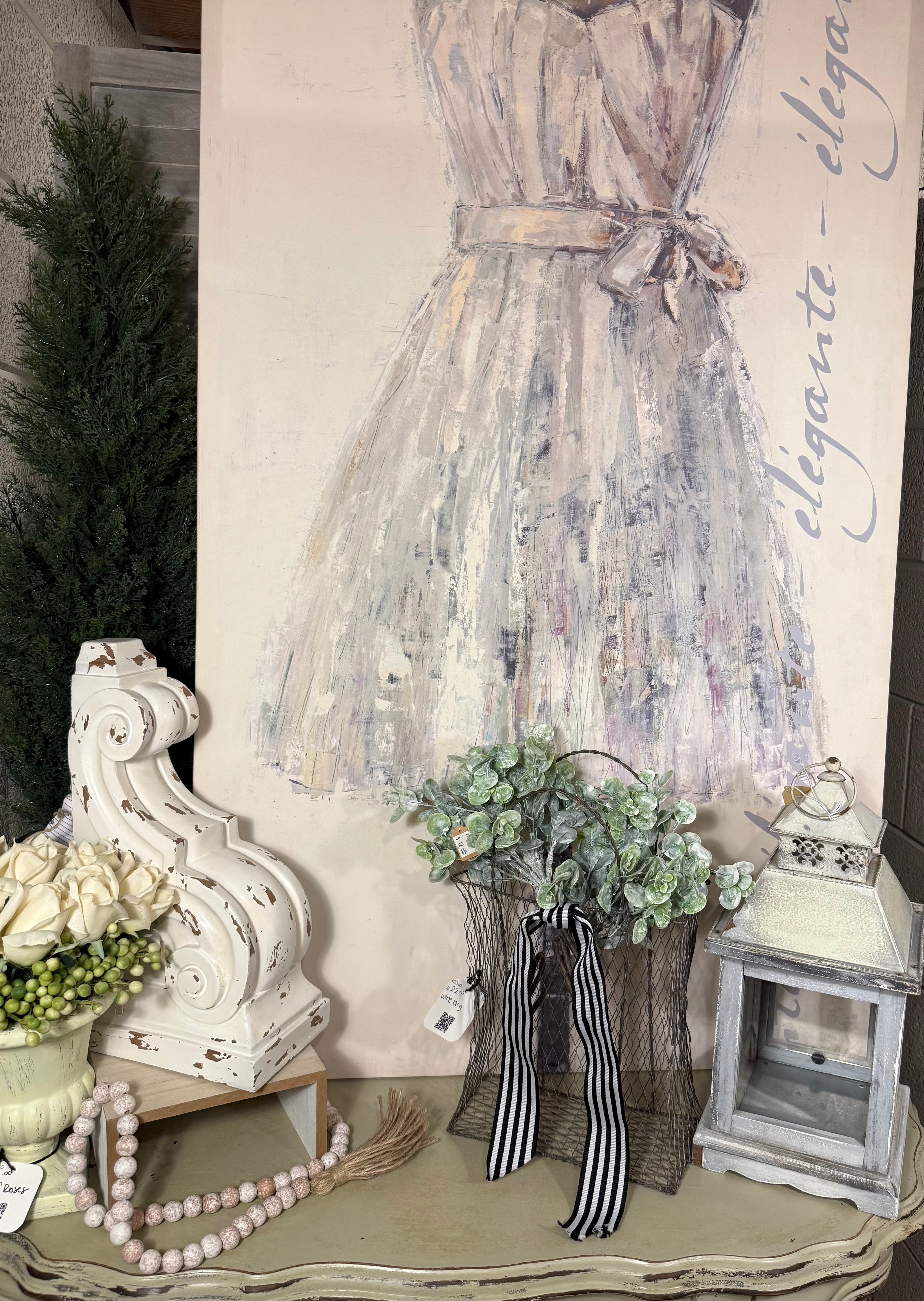 Chic feminine décor setup featuring a pastel dress canvas with 'elegante' script, distressed white corbel, faux white roses, vintage beaded garland, wire basket with greenery and striped ribbon, and a rustic lantern. Perfect for shabby chic, French country, or boutique-style home accents